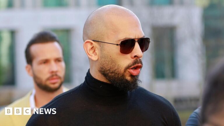 Andrew Tate, a man with a shaved head and dark moustache and beard, faces to the right and speaks, wearing dark glasses and a black turtle-neck shirt