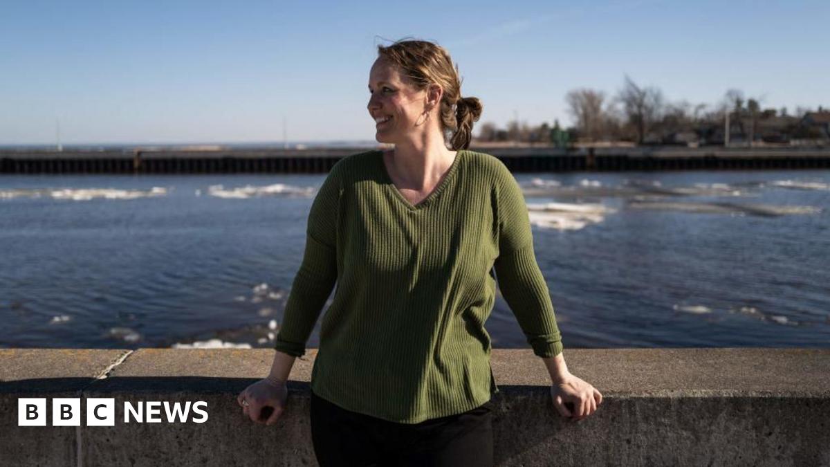 A woman rolls a suitcase to her car parked on an LA street while smoke billows in the distance