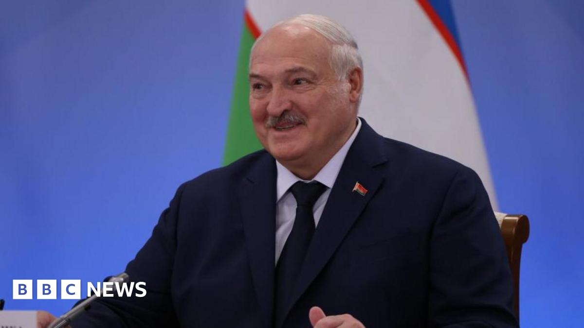 Alexander Lukashenko, sitting at a table with a mic in front of him. He is wearing a dark jacket and tie. A Belarusian flag is in the background.