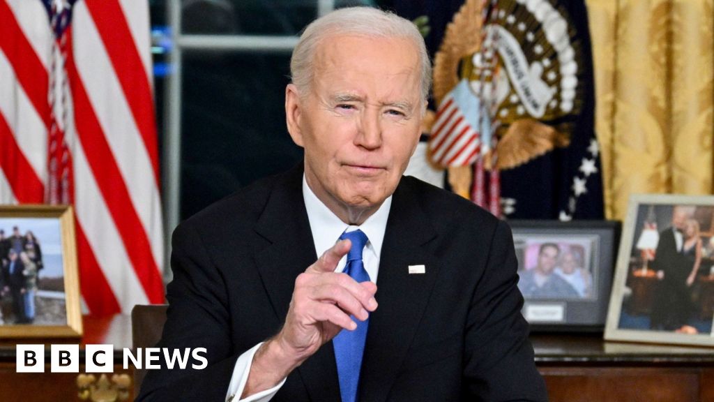 Joe Biden sits at a desk with photos and a US flag behind him and points towards the camera.
