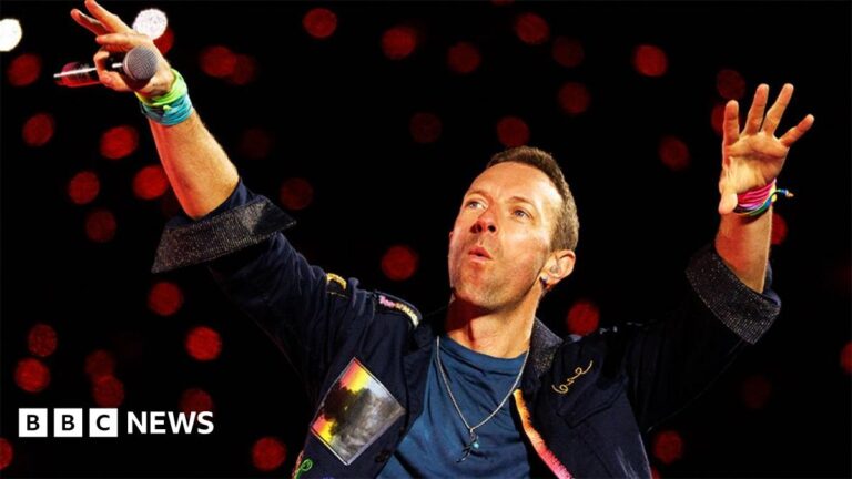Coldplay and Ed Sheeran: Why global stars are now hitting Chris Martin from Coldplay holding both hands up in the air, with a black microphone in his right hand. he is wearing a denim jacket and blue top underneath, with red spotlights behind him.