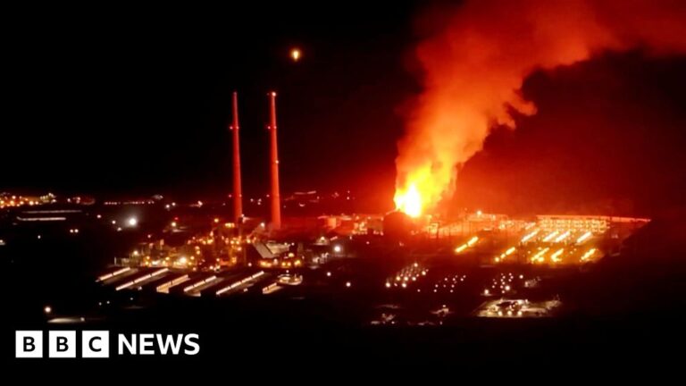 A fire burns at night at a power plant beside two tall chimneys