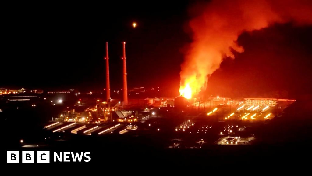 A fire burns at night at a power plant beside two tall chimneys