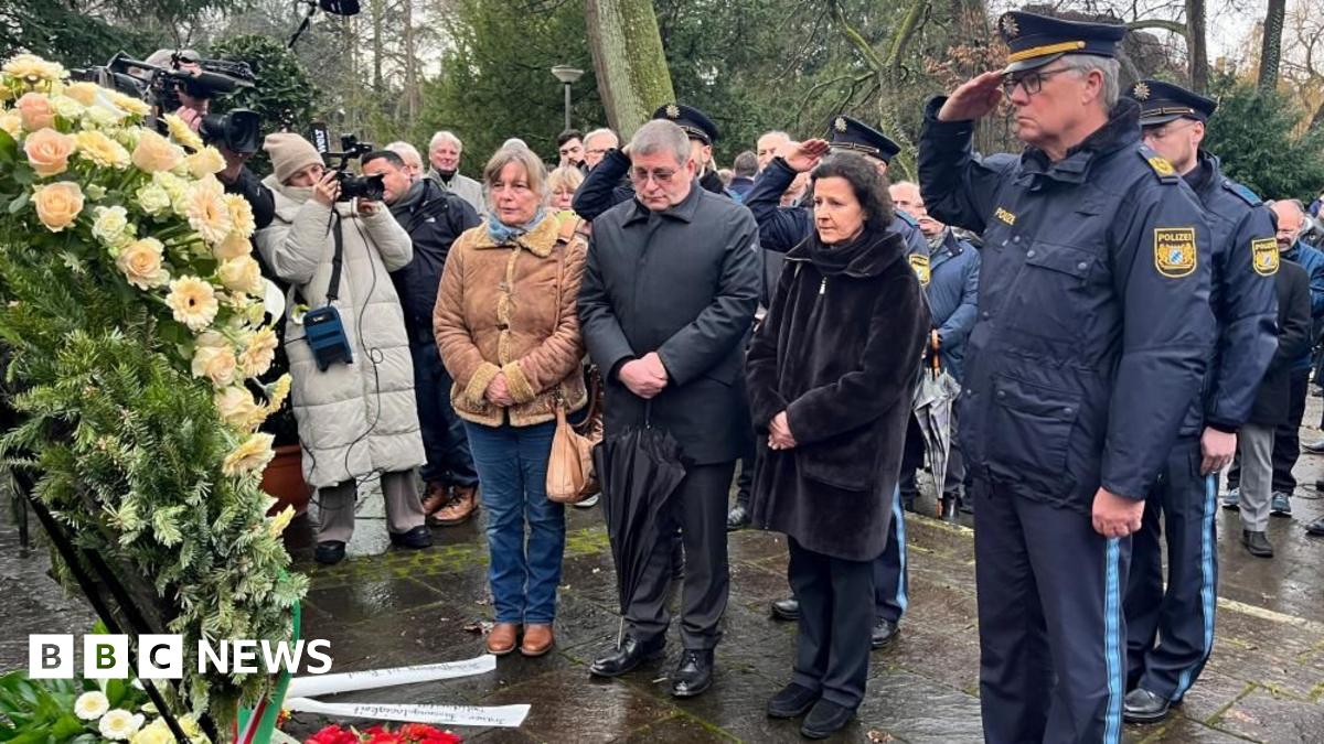 A police officer salutes and other people look on after a wreath of flowers is laid on a rainy day in a park in Bavaria where a toddler and a man were fatally attacked.