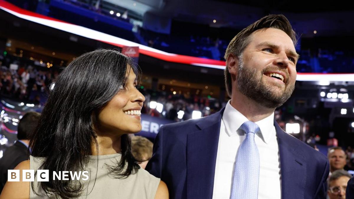 Usha Vance stands with her hands clasped in off-the-shoulder black dress while JD Vance stands next to her in suit and lavender tie, waving his left hand