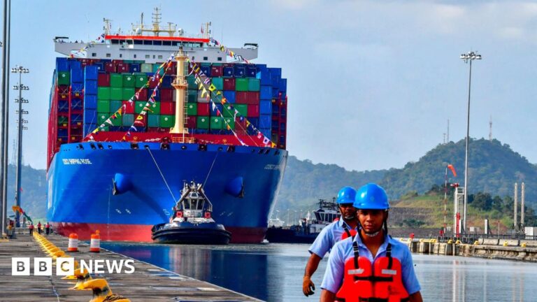 A bright blue container ship sails along the Panama Canal. Two workers in blue helmets and orange hi-vis jackets stand in the foreground.
