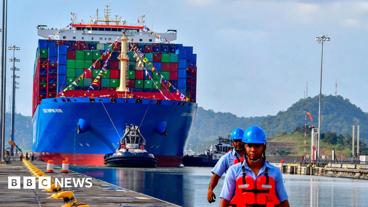 A bright blue container ship sails along the Panama Canal. Two workers in blue helmets and orange hi-vis jackets stand in the foreground.
