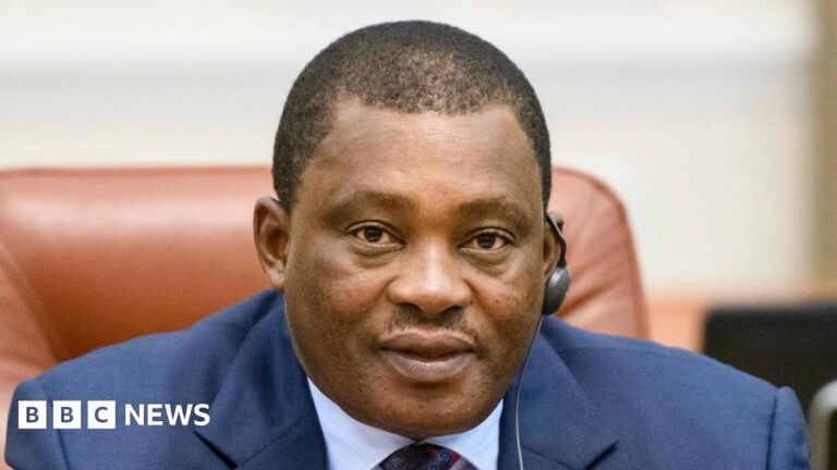 Public Service Cabinet Secretary Justin Muturi speaks in a microphone while seating on a leather chair. He is wearing a navy blue suit jacket and a light blue shirt and a multicoloured tie.