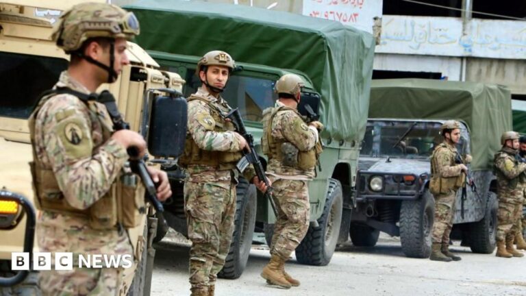 Five Lebanese army soldiers stand with guns in front of three army trucks.
