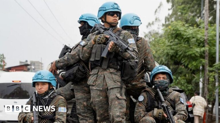 Five soldiers wearing blue hats stand on the back of a truck in front of trees in Goma, DR Congo.