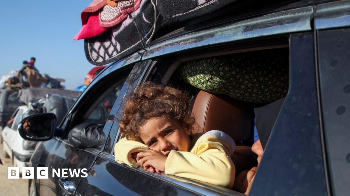 A young girl, wearing a yellow jumper leans her cheek on her arms crossed in the open window of a car that is piled with belongings, other vehicles visible ahead of it, as Gazans wait to return to the north of the territory