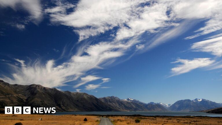 The image shows blue skies and mountains, with a country road leading to Lake Ohau, a glacial lake in the Mackenzie Basin of the South Island in New Zealand.