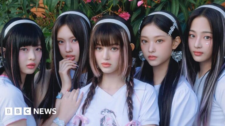 Promotional photo of the band NewJeans, with the five members dressed in white t-shirts, with white alice bands in their hair, against a floral backdrop.