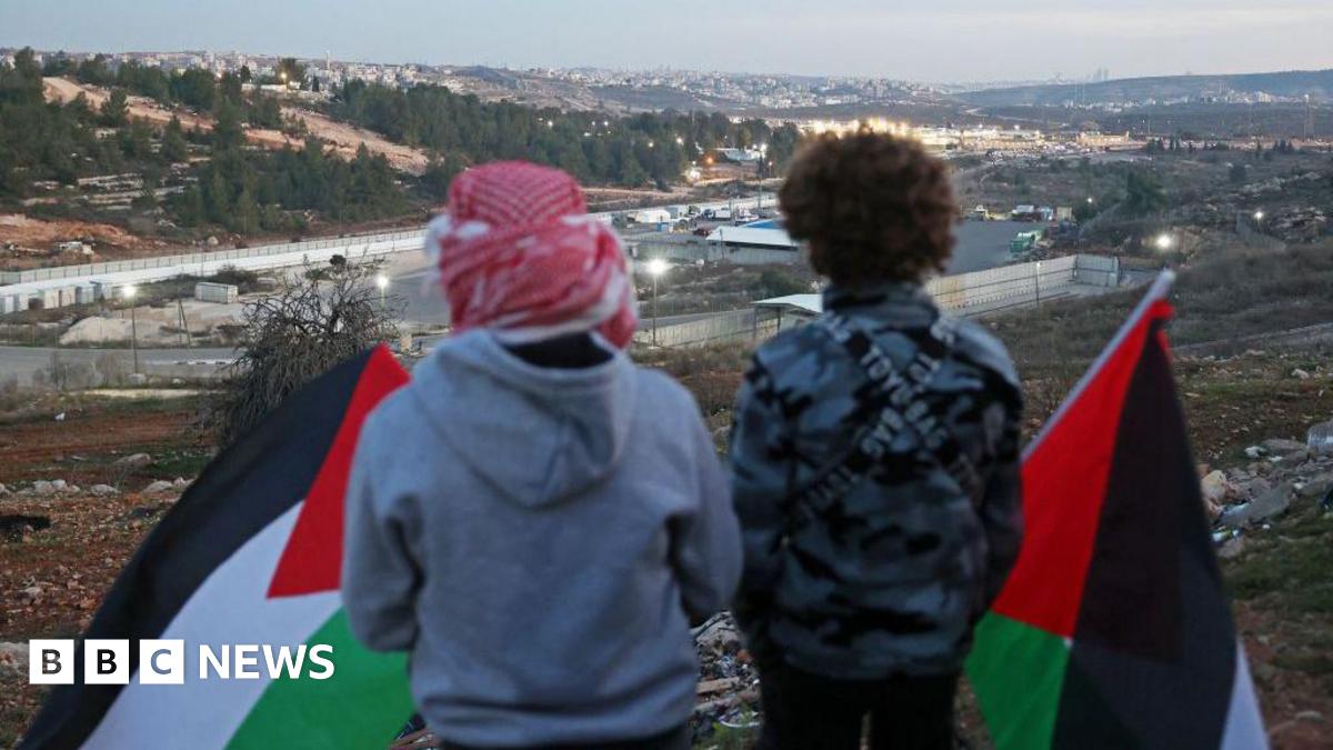 Two Palestinians with their backs to the camera hold black, white, green and red Palestinian flags while look down on Ofer prison, in the occupied West Bank, ahead of the release of dozens of Palestinian prisoners in exchange for three Israeli hostages on the first day of the Gaza ceasefire deal (19 January 2025)