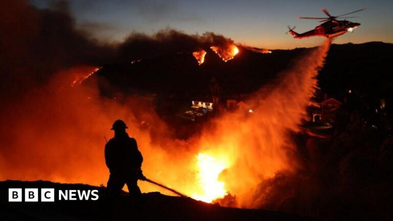 The silhouette of a firefighter is seen against bright orange flames as a helicopter drops water on burning hills in the distance