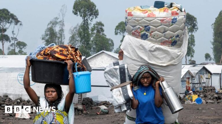 Two women flee a camp for displaced people with their belongings. They carry a mattress, a large thermos flash and more.