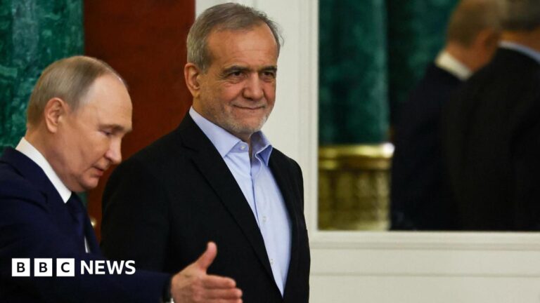 Vladimir Putin, seen in profile, gestures towards his Iranian counterpart, Masoud Pezeshkian, who is walking slightly ahead of him. Both men are wearing dark suits. Putin has a white shirt and dark tie, while Pezeshkian wears a blue open-necked shirt.