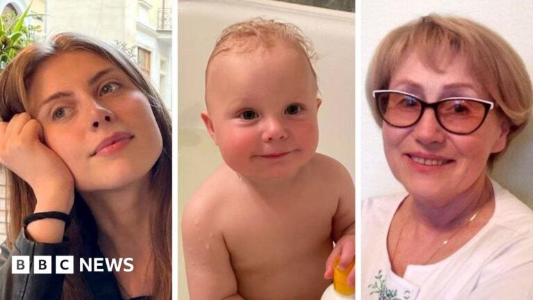 A lady with short blonde hair looks into the camera. Behind her is her apartment which has been totally demolished by a bomb.