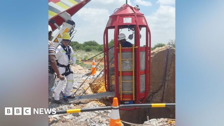 A man in a white boiler stands by a crane that is about to lower a cage down a mine shaft.