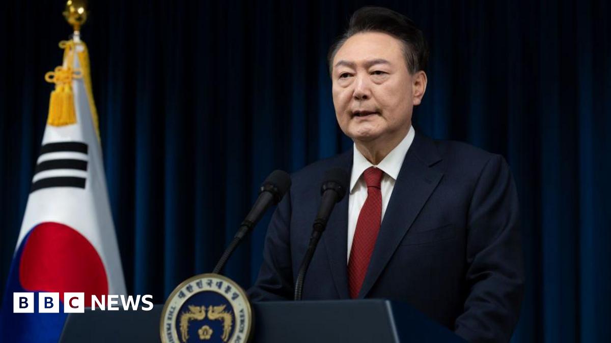 Yoon Suk Yeol wearing a navy suit and red tie speaking at a podium. A large South Korean flag is beside him.