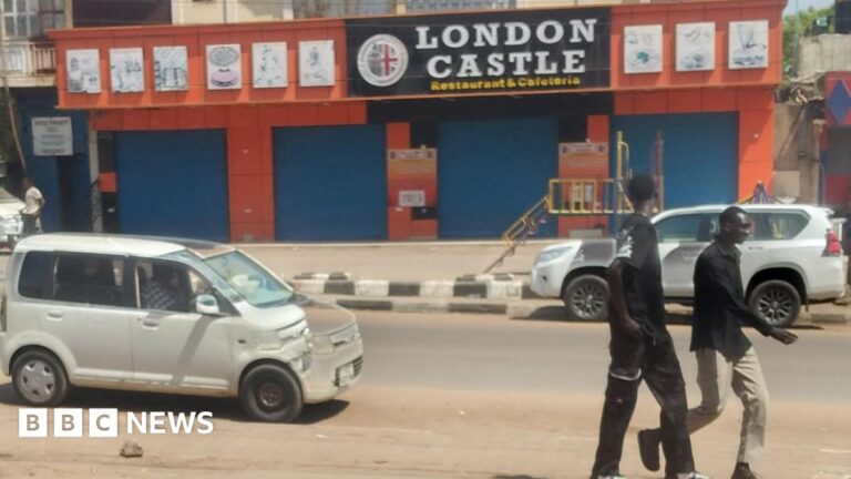 Two men walk past a shuttered restaurant and parked cars on a quite street in Juba during the daytime. The restaurant is called