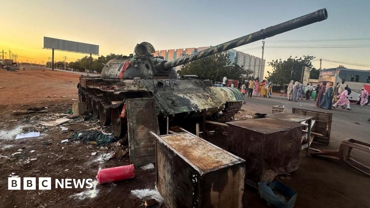 A military tank is standing on the side of a road in Wad Mdani, with people in civilian clothes nearby