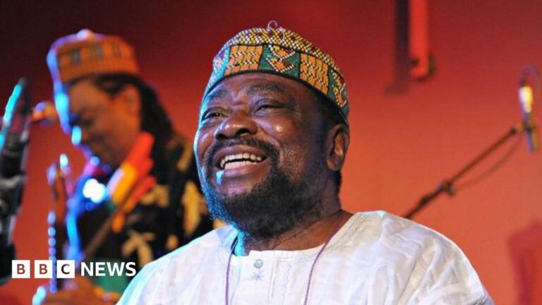 Teddy Osei, dressed in a crisp white linen shirt and a traditional African cap, radiates joy with a broad smile as he performs with his band Osibisa at the Front Room of the Queen Elizabeth Hall in 2010.