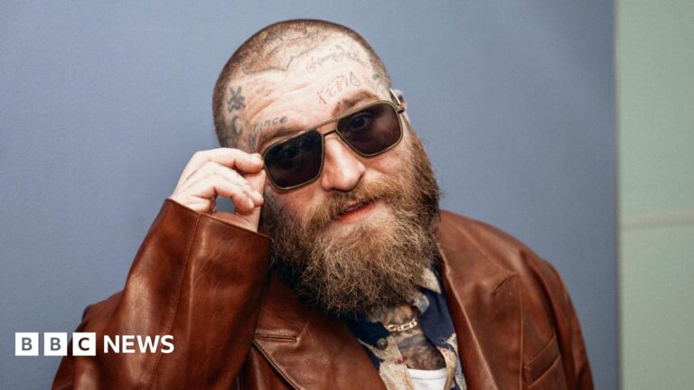 Teddy Swims, wearing a brown leather jacket, tips his sunglasses towards the camera as he poses at the BBC