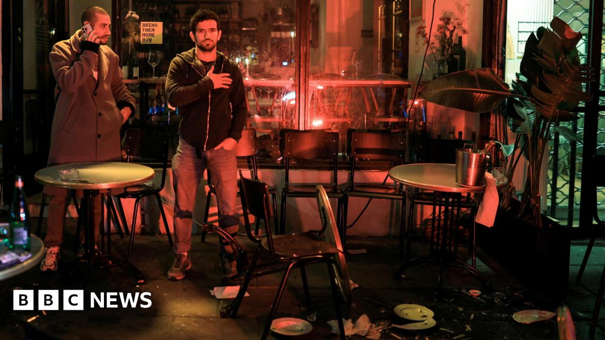 People stand outside a wine bar near the scene of a suspected attack in Tel Aviv. One man faces the camera with a hand on his chest. Cracked china plates and splintered glass lie on the ground and a table is upturned.
