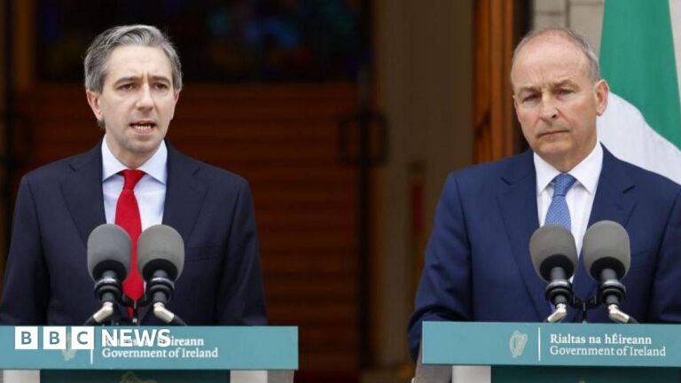 Simon Harris and Micheál Martin both stand in front of green Government of Ireland branded podiums. Mr Harris is wearing a navy suit, light blue shirt and red tie. Mr Martin is wearing a dark blue suit, white shirt and dark blue tie.