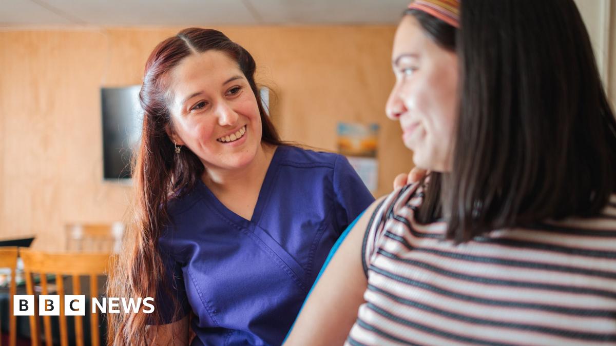 Two women smiling at each other. One is a medical practitioner and is wearing blue scrubs, the other is a patient who seems to be being examined.