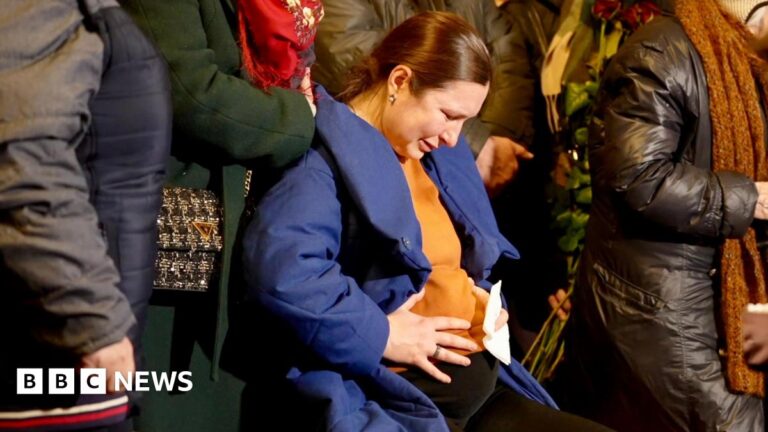 Anastasiia sits looking down sad as her hands rest on her stomach, surrounded by others standing