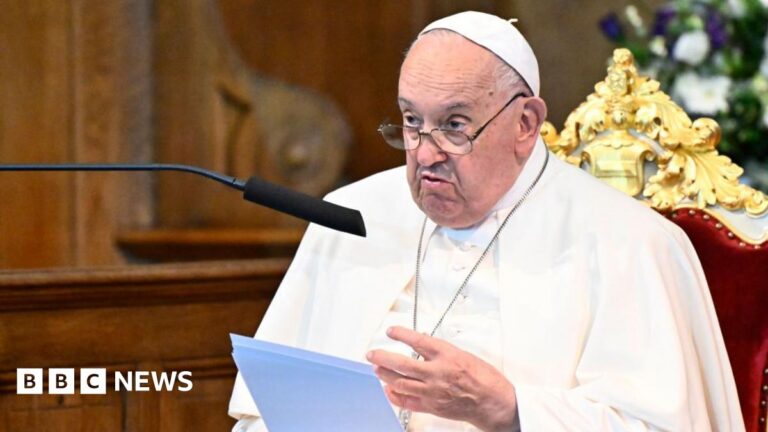 Pope Francis sitting in a velvet chair, reading from a sheet of paper. He is speaking into a microphone. The background is blurred.