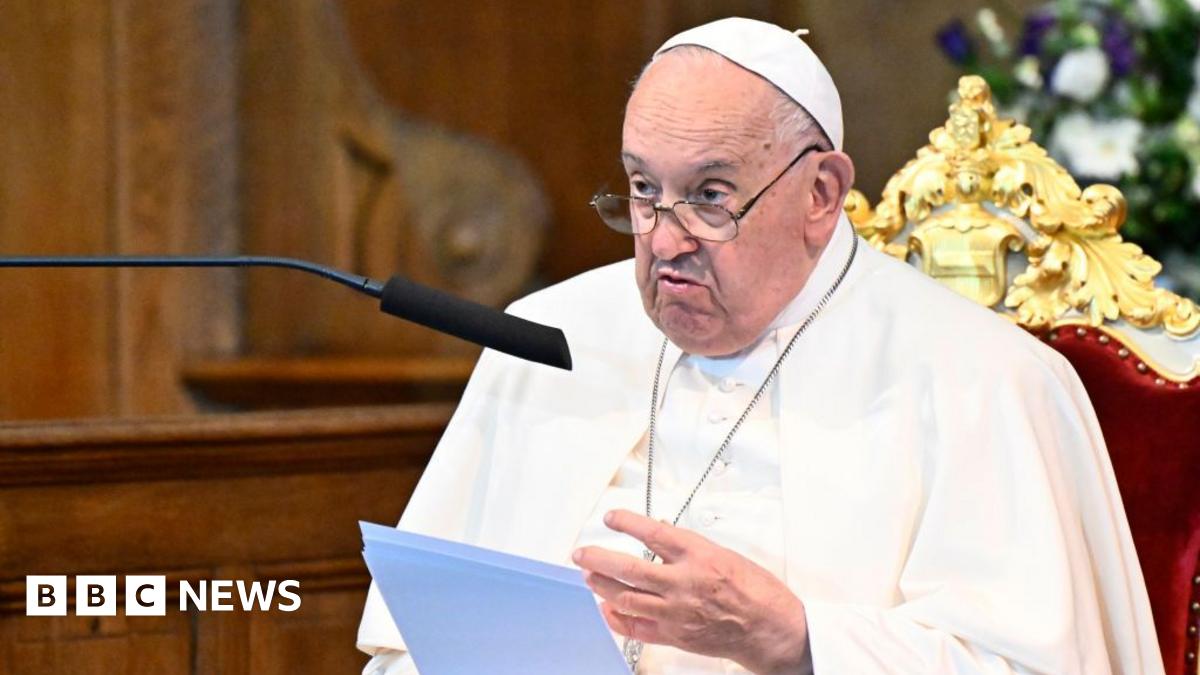 Pope Francis sitting in a velvet chair, reading from a sheet of paper. He is speaking into a microphone. The background is blurred.