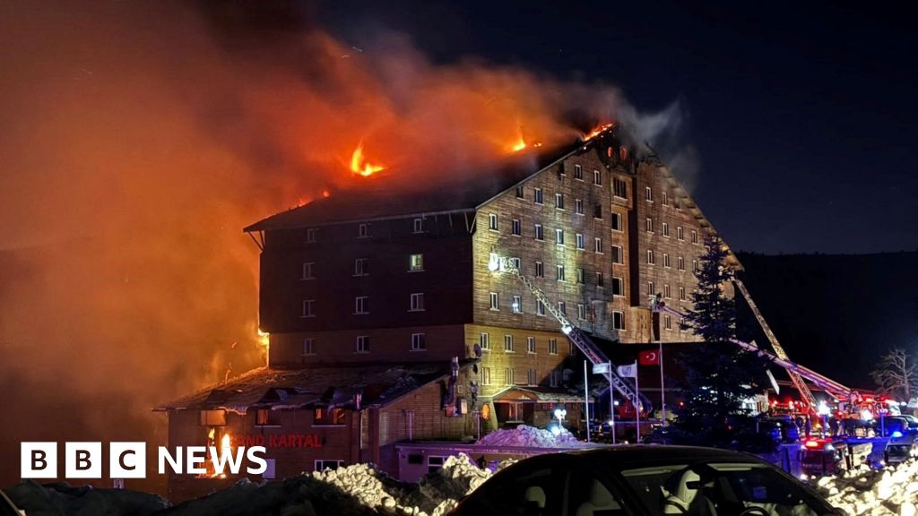 The exterior of the Grand Kartal Hotel at night with the roof in flames and two ladders from fire trucks extended.