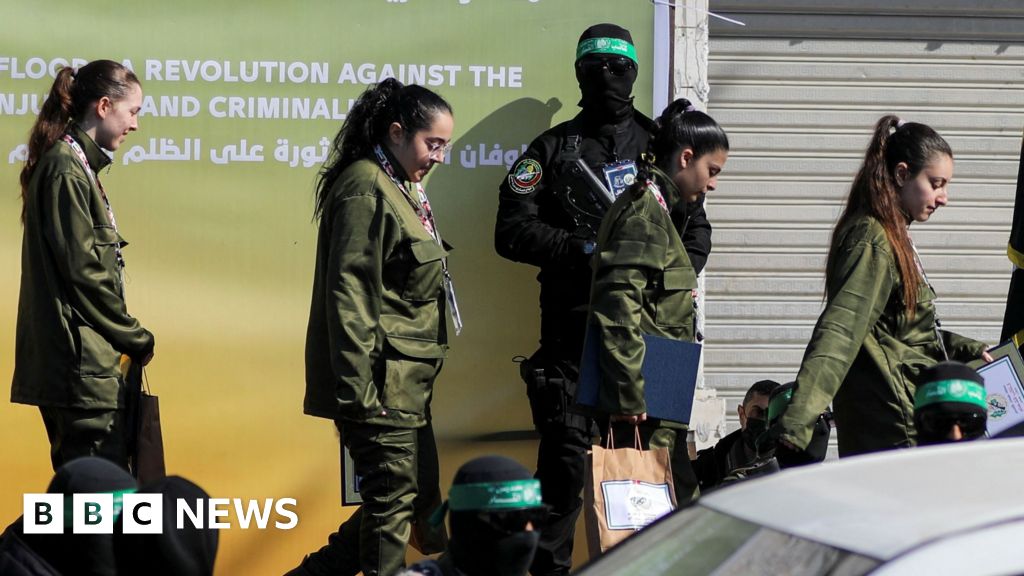 Four female Israeli soldiers in army uniform walking in a line, behind them is an armed Hamas fighter
