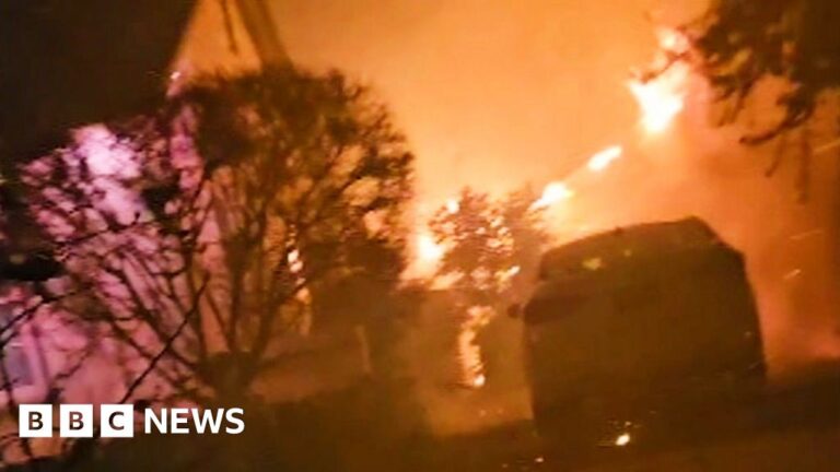 A blazing fire is seen engulfing a garage with a car parked in front of it