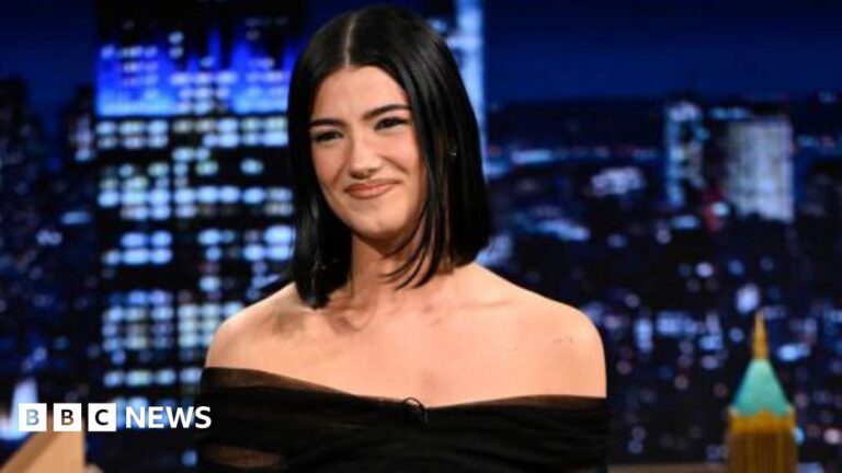 A woman with straight black hair smiles. She is in the studio of the Jimmy Fallon show.