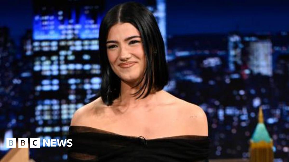 A woman with straight black hair smiles. She is in the studio of the Jimmy Fallon show.