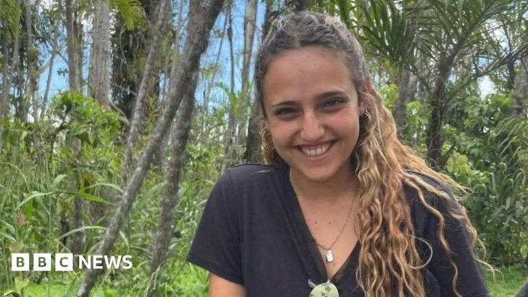File image of Romi Gonen smiling at the camera. She has curly brown hair and is wearing a black top